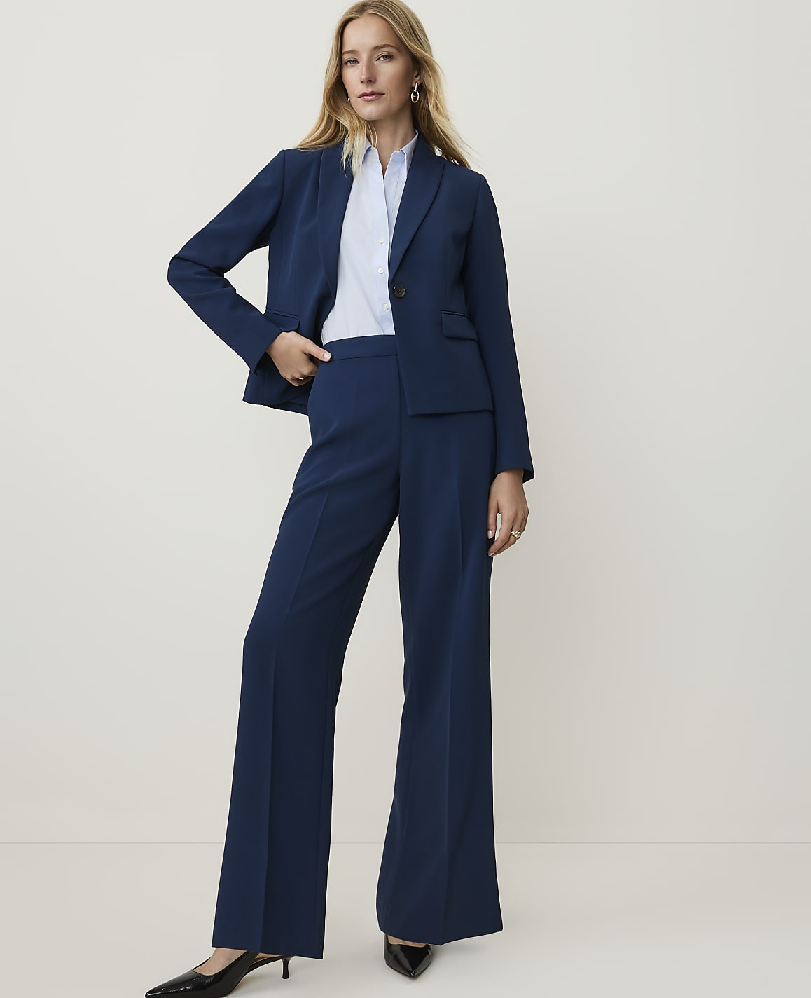 A woman in a navy blue pantsuit stands against a plain, light background.