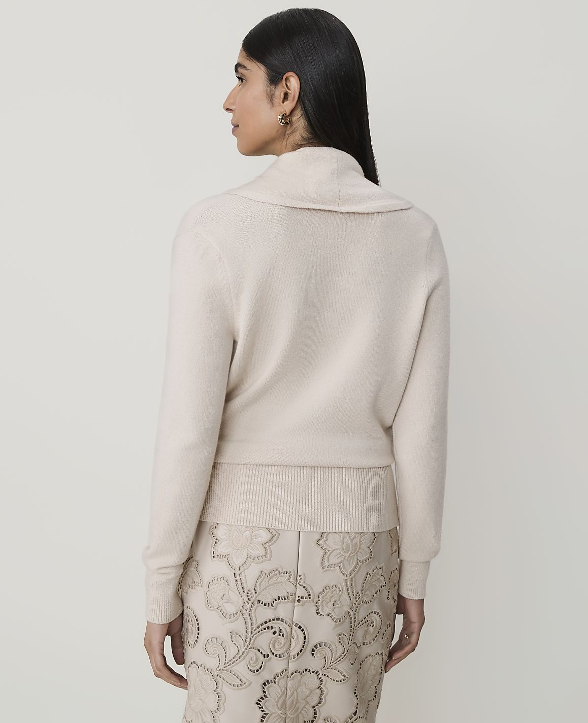 A woman in a cream cowl-neck sweater and a patterned skirt stands against a plain, light background.