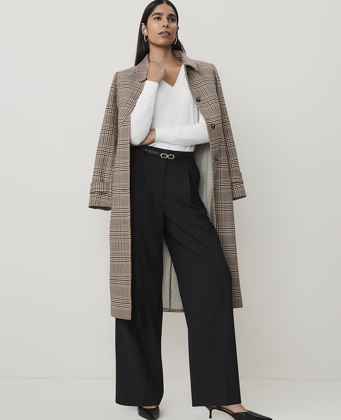 A woman in a white top, black wide-leg pants, and a plaid coat stands against a plain, light-colored background.
