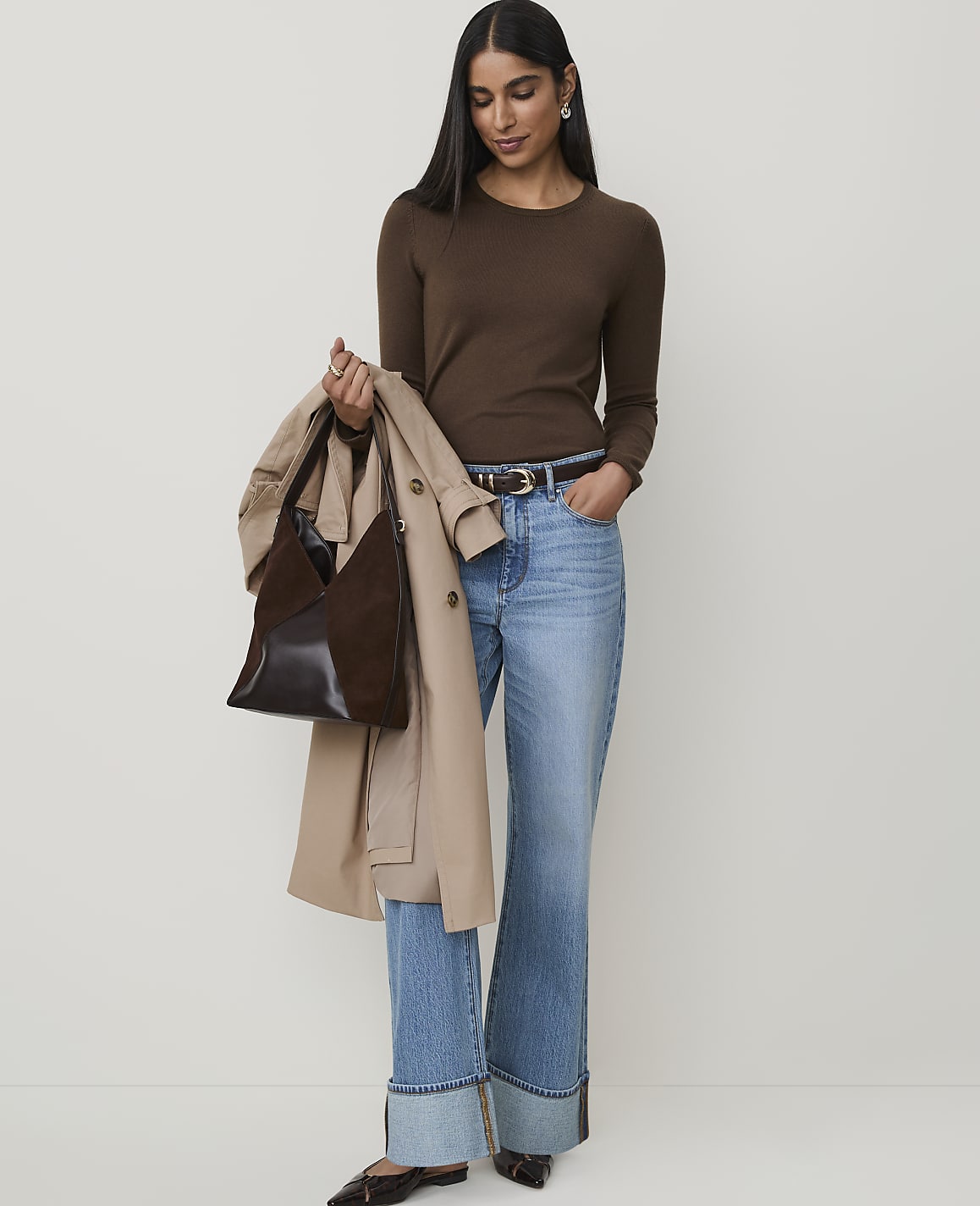 A woman in a brown long-sleeved shirt and flared jeans stands against a plain white background, holding a trench coat and a dark brown handbag.
