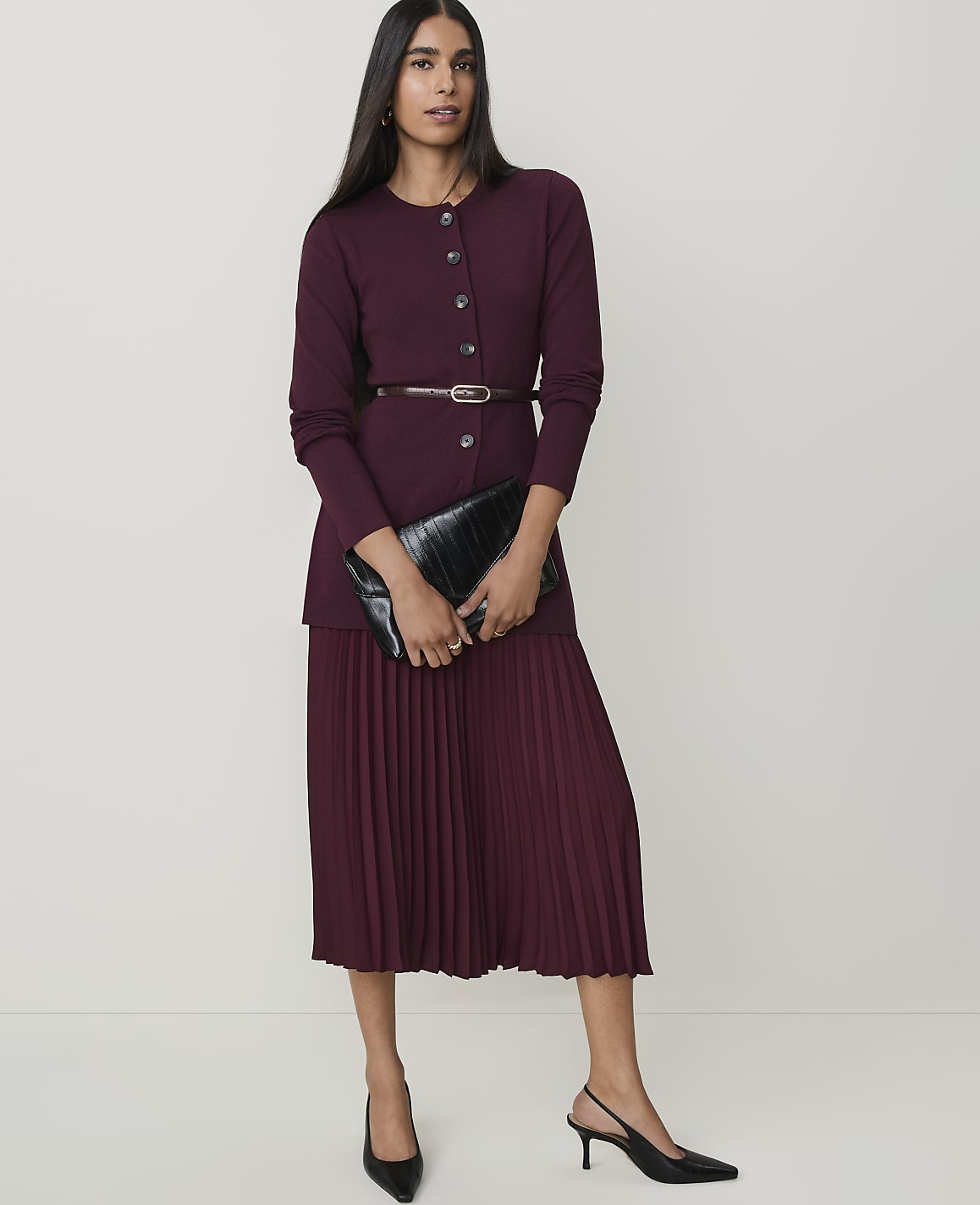 A woman in a burgundy cardigan and pleated skirt stands against a plain white background, holding a black clutch.