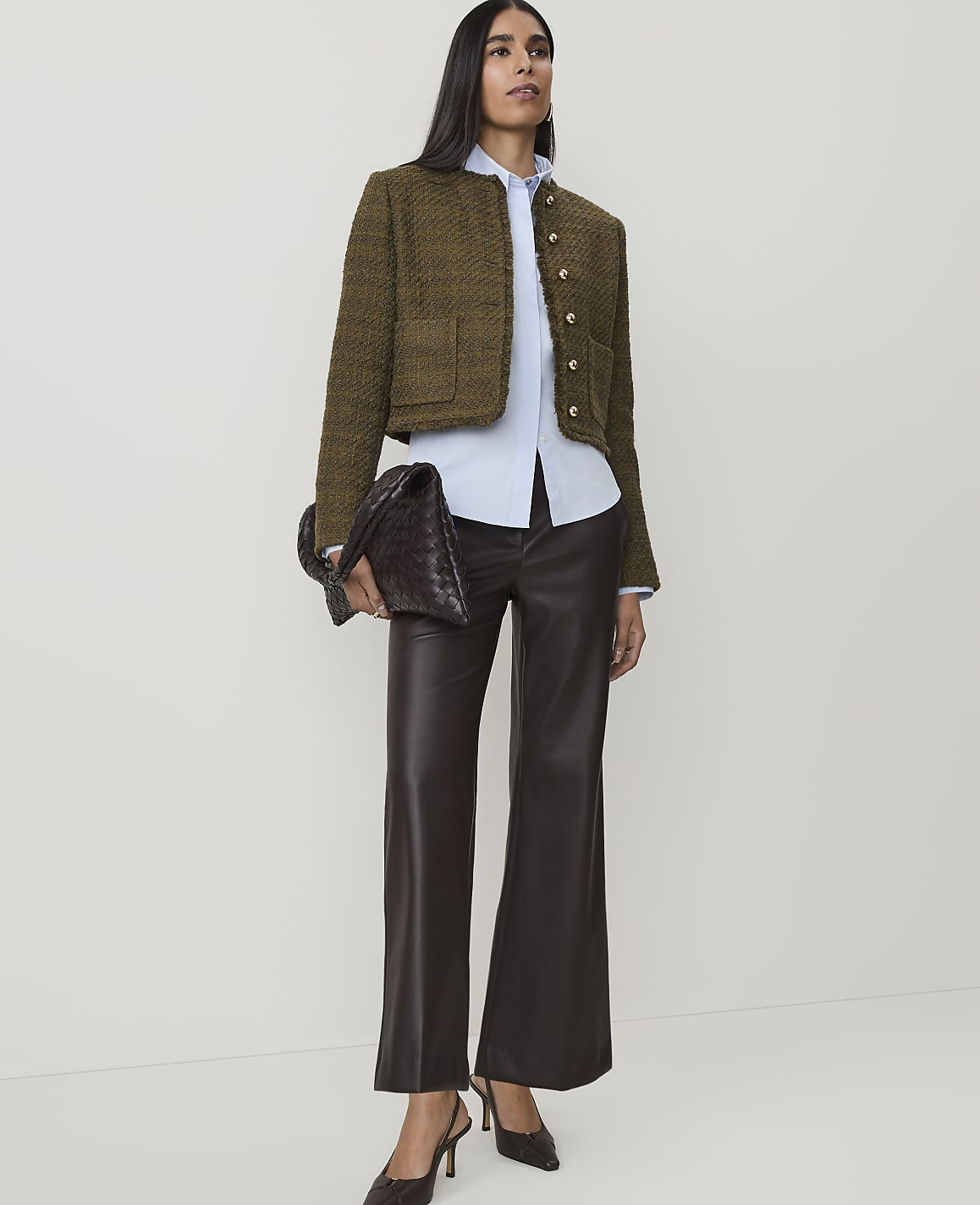 A woman in a tweed jacket and leather pants stands against a plain white background, holding a woven clutch.