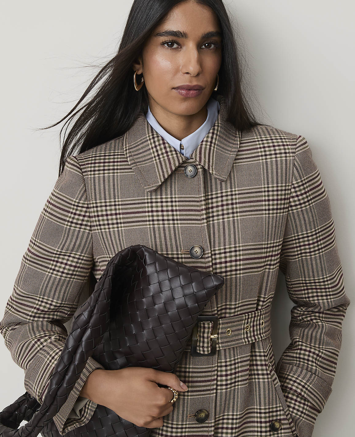 A woman in a plaid coat holds a woven bag in front of a plain background.