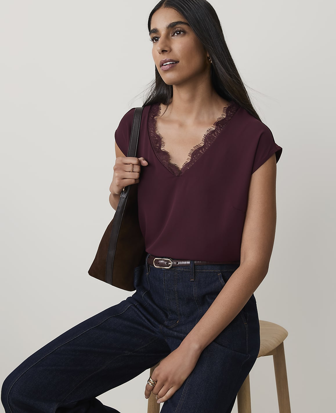 A woman in a burgundy lace-trimmed top and dark jeans sits against a plain, light background, carrying a brown bag over her shoulder.