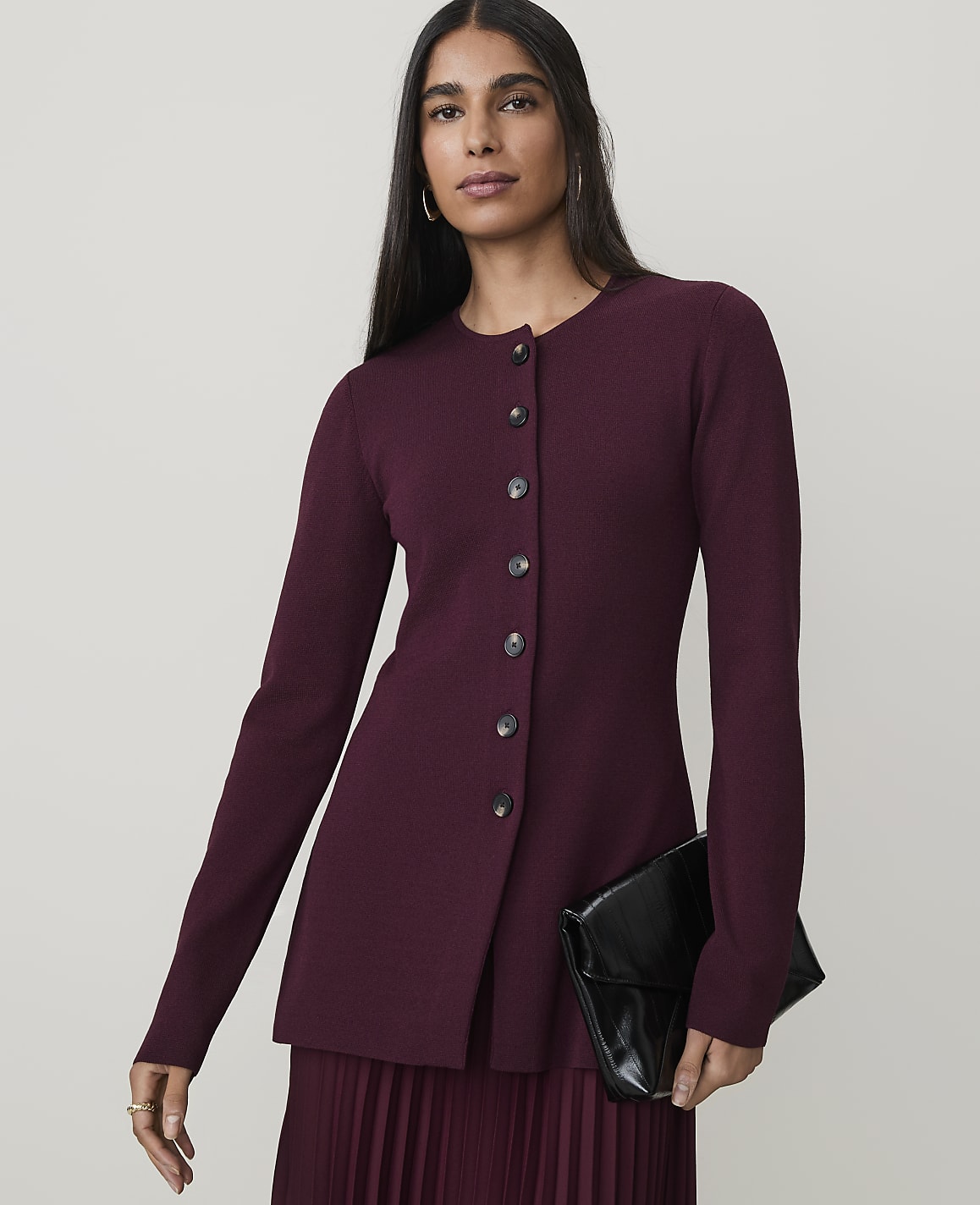 A woman in a burgundy cardigan and pleated skirt holds a black clutch against a plain background.
