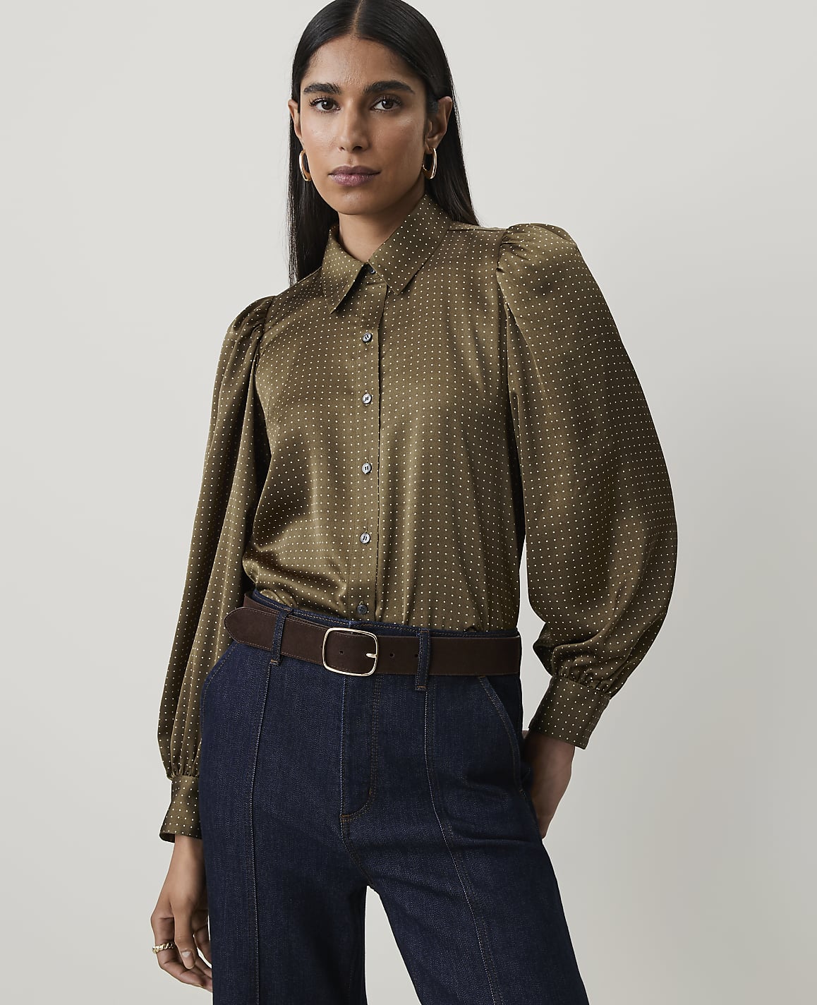 A woman in a patterned olive green blouse and dark jeans stands against a plain, light gray background.