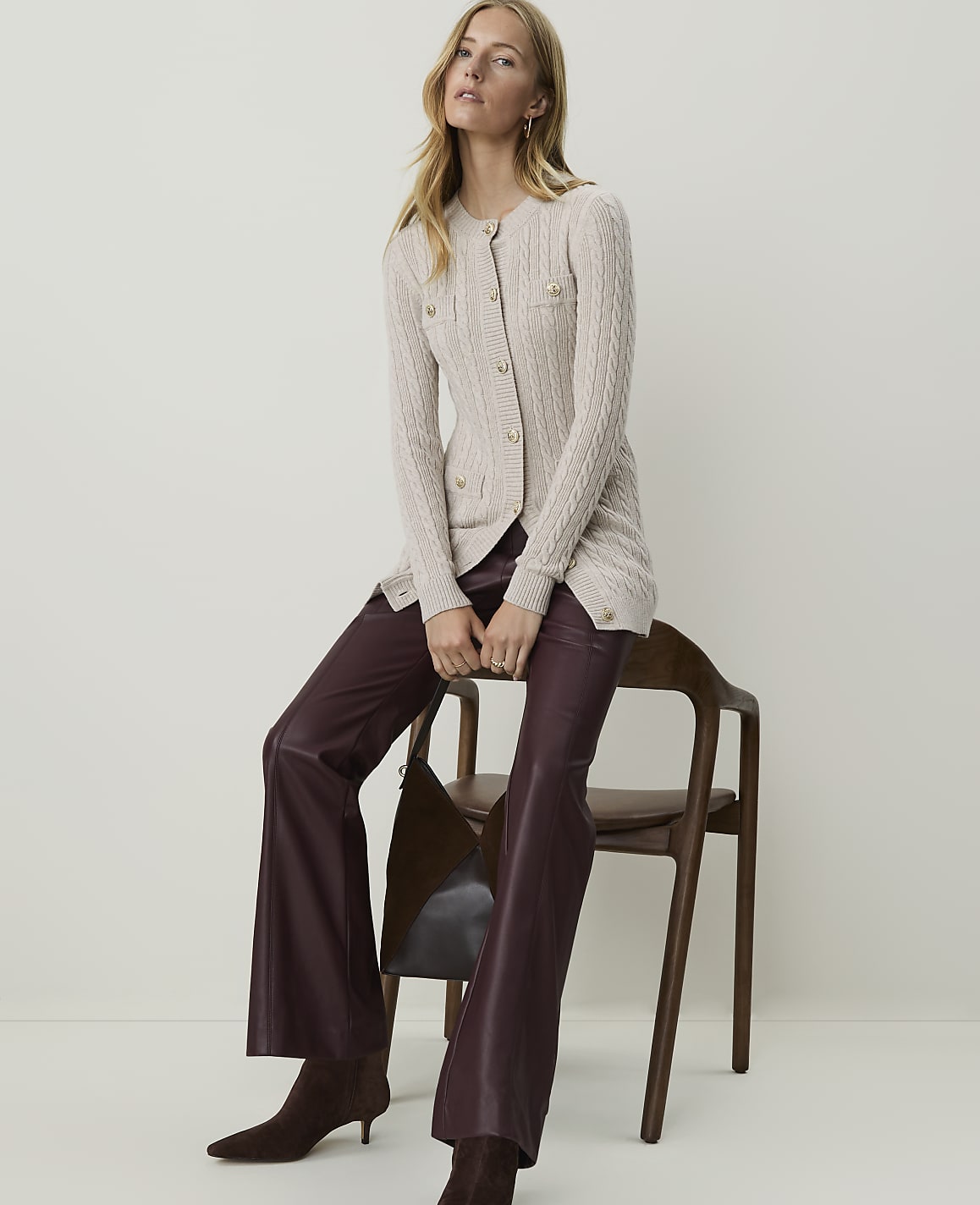 A woman in a cream cable-knit cardigan and burgundy leather pants sits on a wooden chair against a plain, light-colored background.
