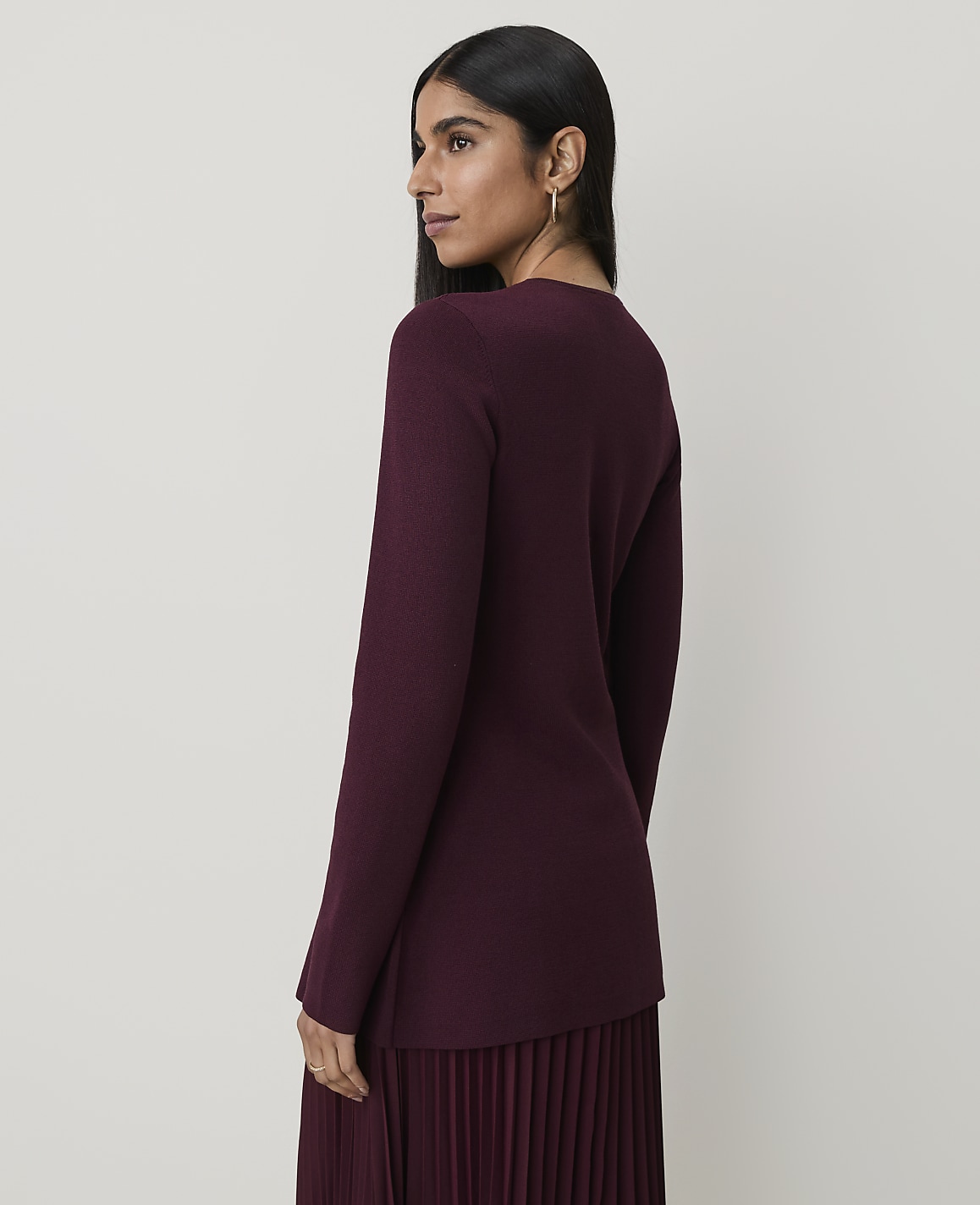 A woman in a burgundy sweater and pleated skirt stands against a plain, light-colored background.