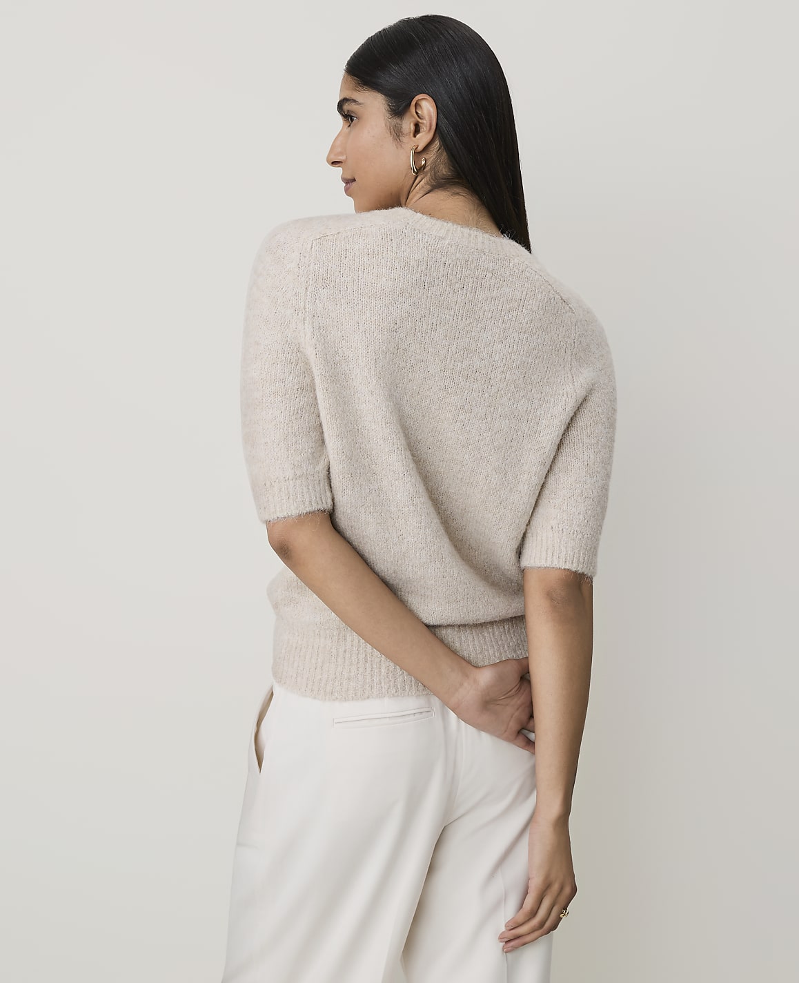 A woman in a cream sweater and pants stands against a plain, light background.