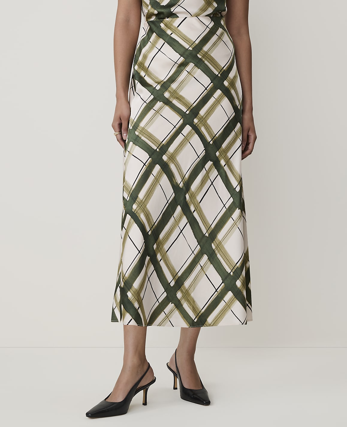 A woman in a cream and green plaid skirt and black slingback heels stands against a plain white background.