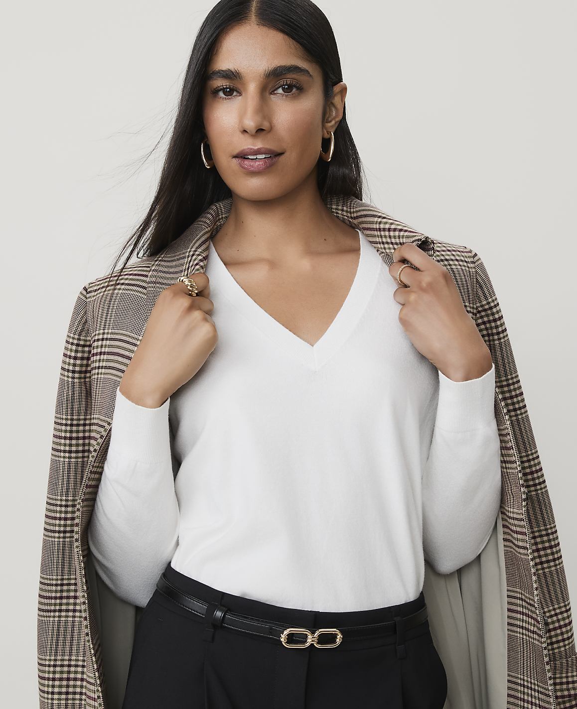 A woman in a white v-neck sweater and plaid blazer poses against a plain background.