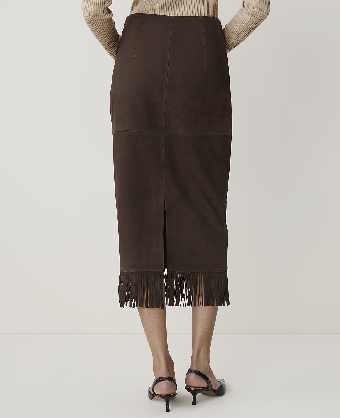 A person stands in front of a plain background, wearing a brown suede skirt with fringe at the hem and black heeled shoes.