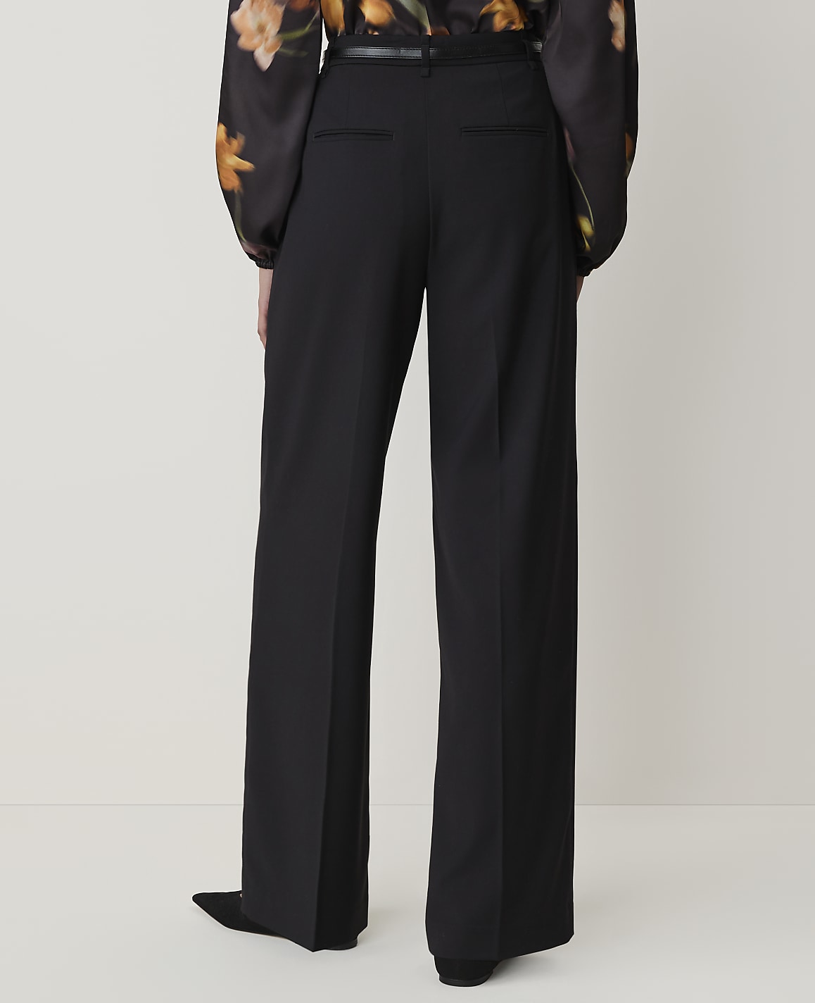 A person wearing black wide-leg pants and a floral blouse stands against a plain, light-colored background.
