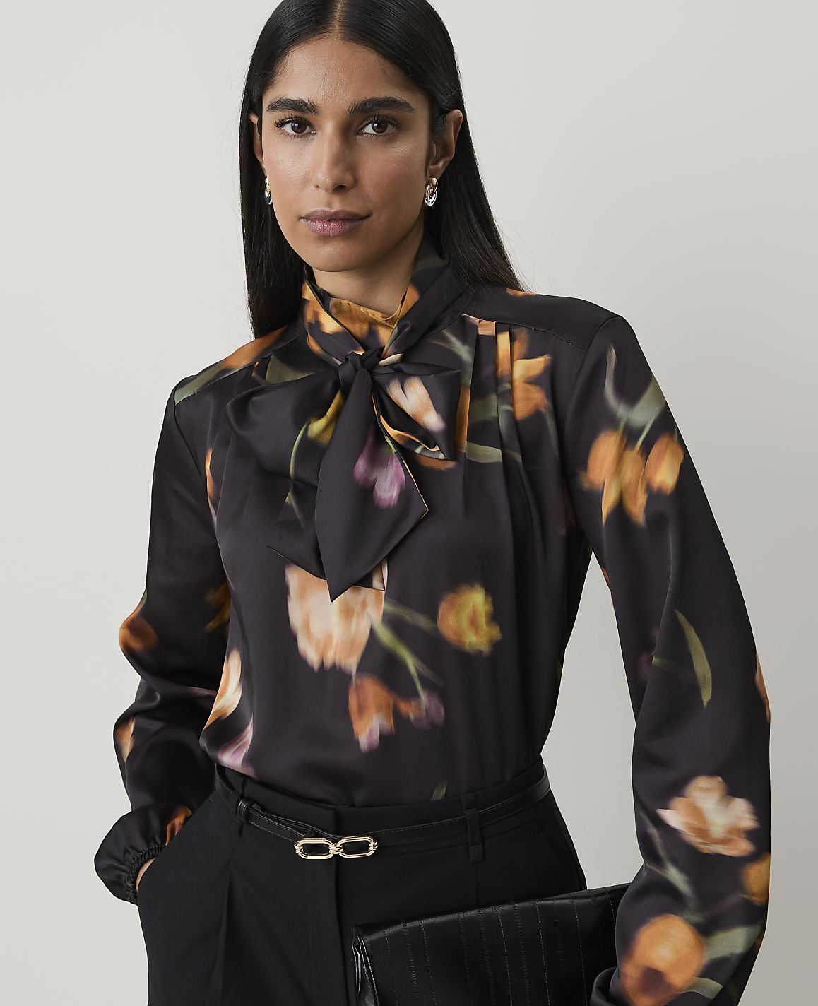A woman in a floral print blouse and black pants holds a clutch in front of a plain background.