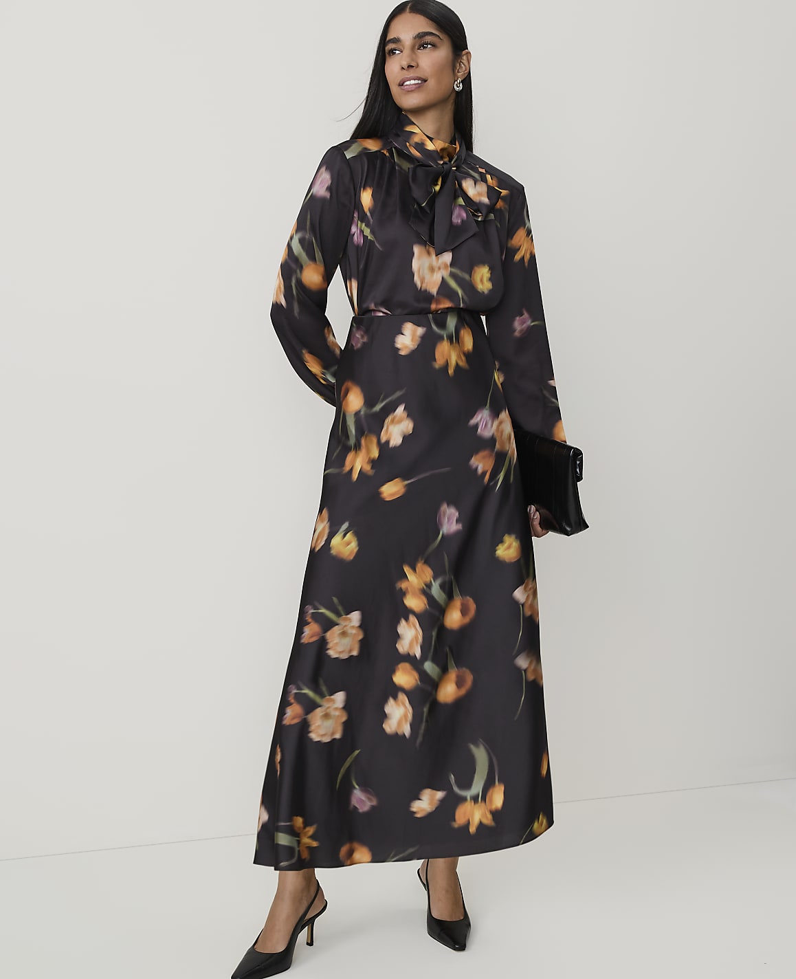 A woman in a floral dress stands against a plain white background, holding a black clutch.