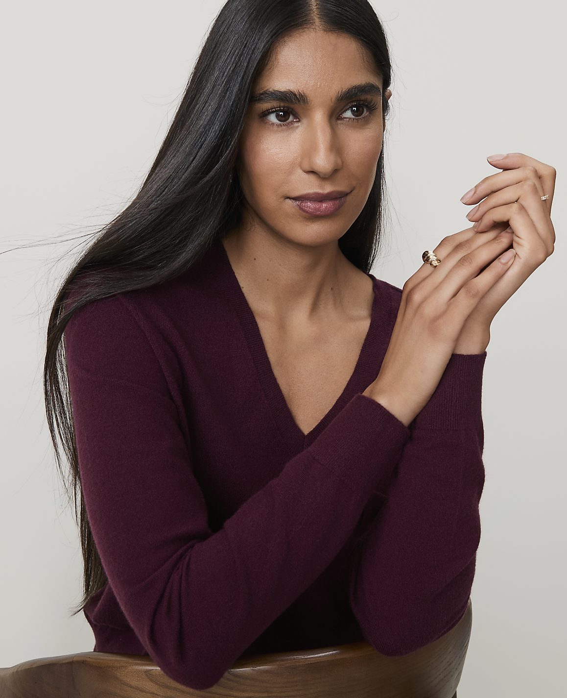 A woman with long dark hair and a burgundy v-neck sweater rests her chin on her hands, with a blurred wooden chair in the foreground and a plain background.