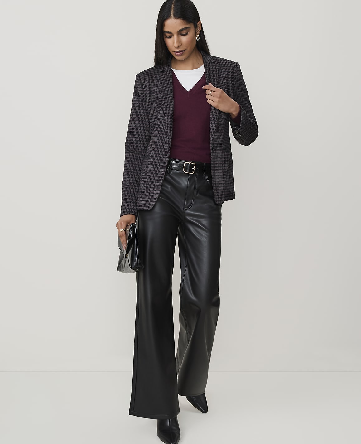 A woman in a patterned blazer, burgundy v-neck sweater, and black leather pants stands against a plain white background, holding a black clutch.