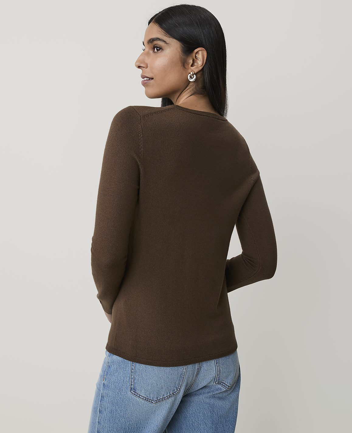 A woman in a brown sweater and blue jeans stands against a plain, light-colored background.