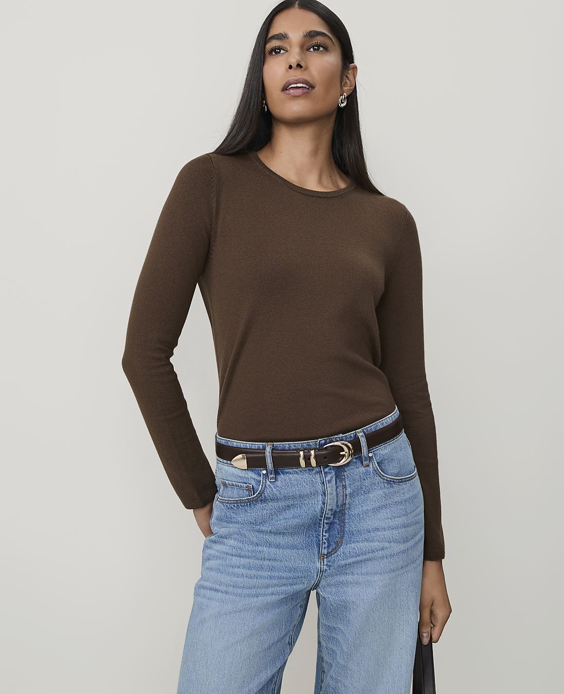 A woman in a brown long-sleeved shirt and light blue jeans stands against a plain, light gray background.