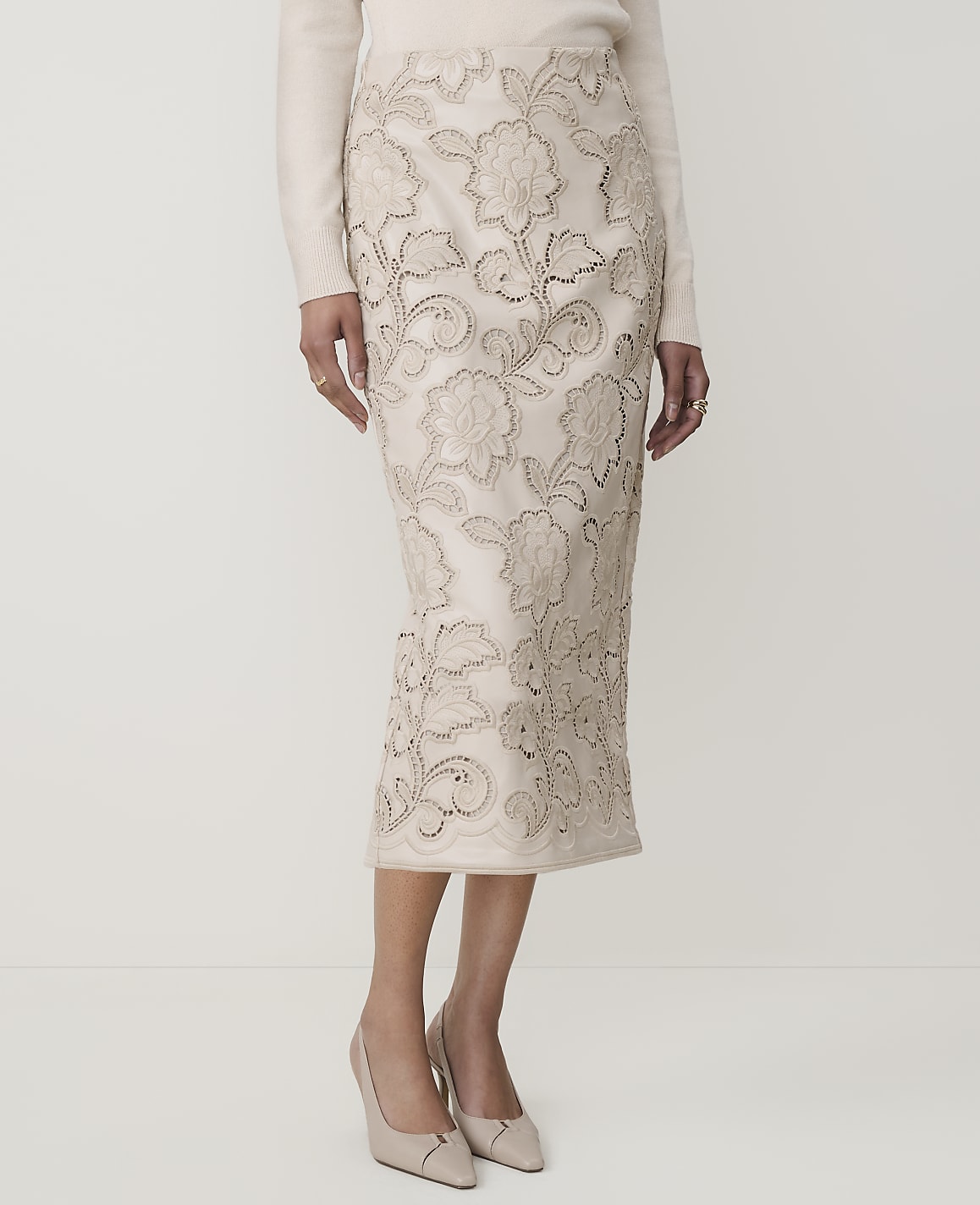 A woman models a cream-colored, floral-embroidered pencil skirt and matching heels against a plain white background.