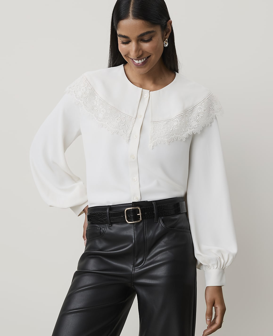 A woman in a white lace-trimmed blouse and black leather pants smiles against a plain background.