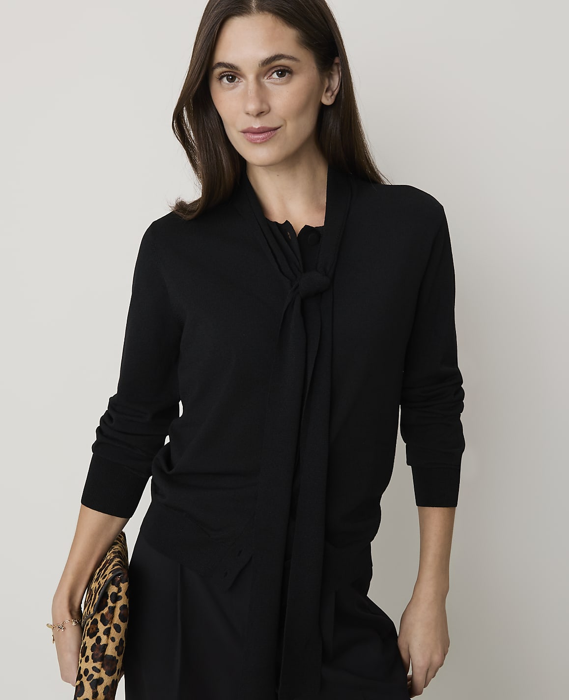 A woman in a black sweater with a bow detail holds a leopard-print clutch against a plain background.