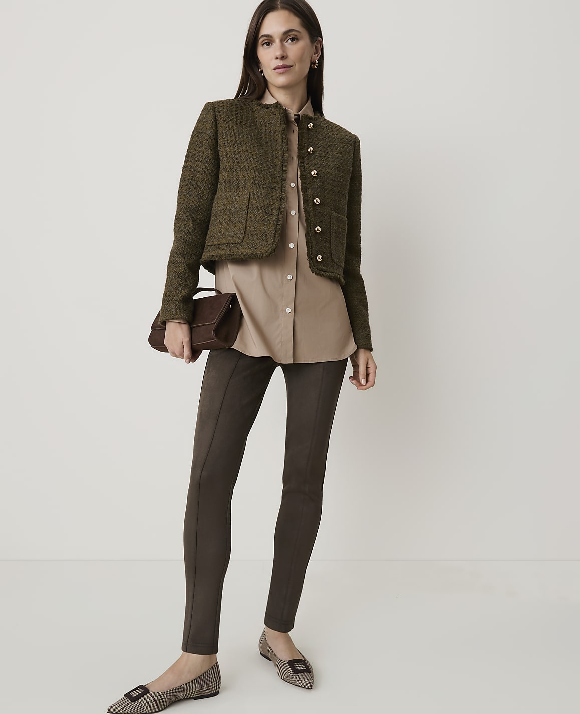 A woman in a tweed jacket and leggings stands against a plain, light background, holding a clutch.