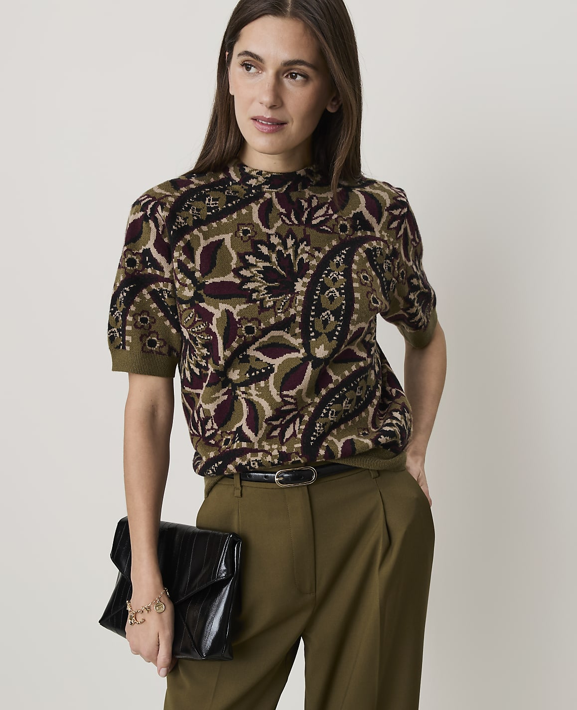 A woman in a patterned top and olive pants holds a black clutch in front of a plain background.