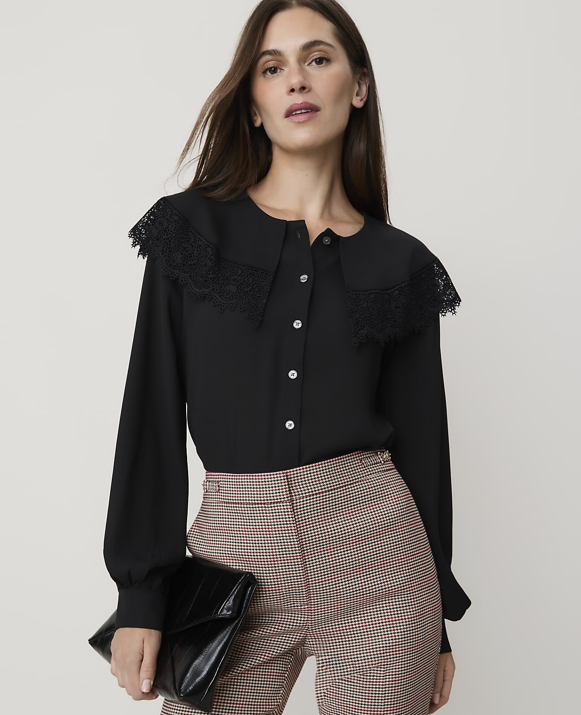 A woman in a black blouse and plaid pants holds a black clutch in front of a plain background.