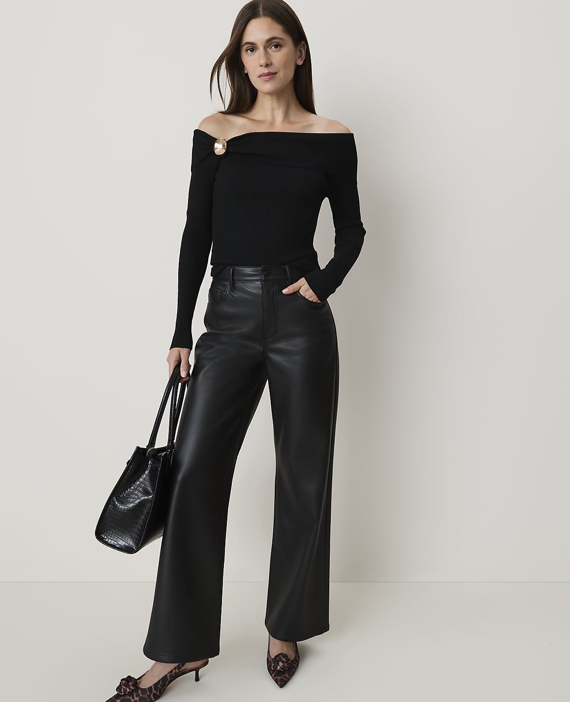 A woman in a black off-the-shoulder top and leather pants stands against a plain white background, holding a black handbag.