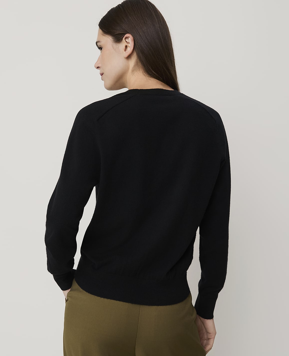 A woman in a black sweater and olive pants stands against a plain, light-colored background.