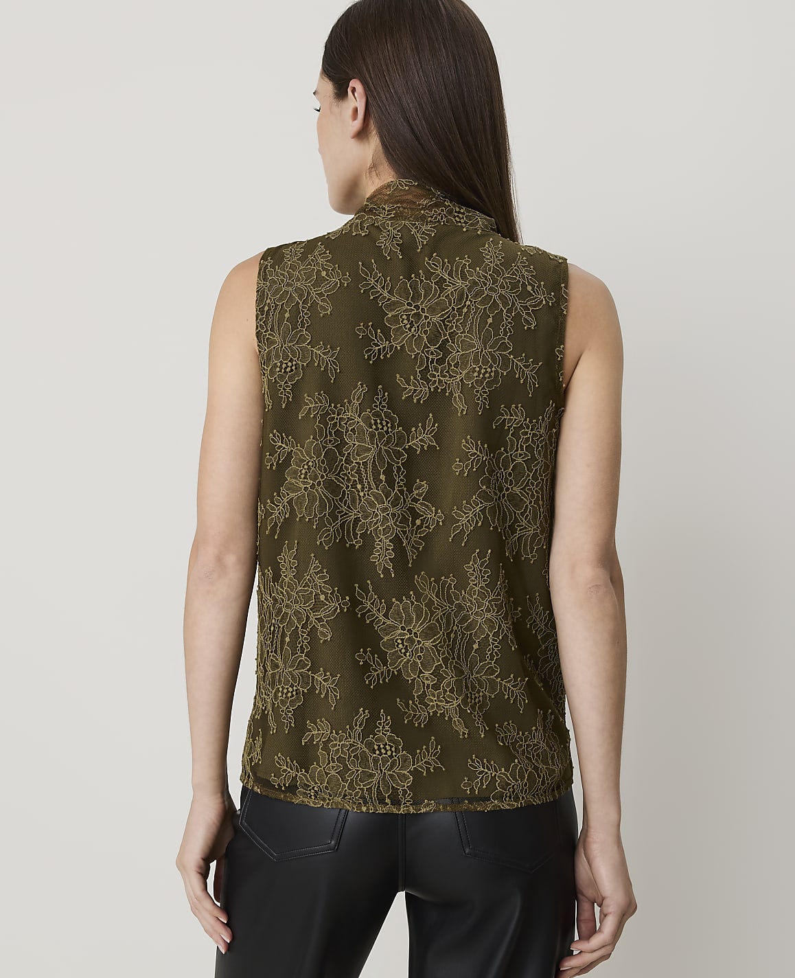 A woman in a sleeveless olive green lace top and black pants stands against a plain white background.