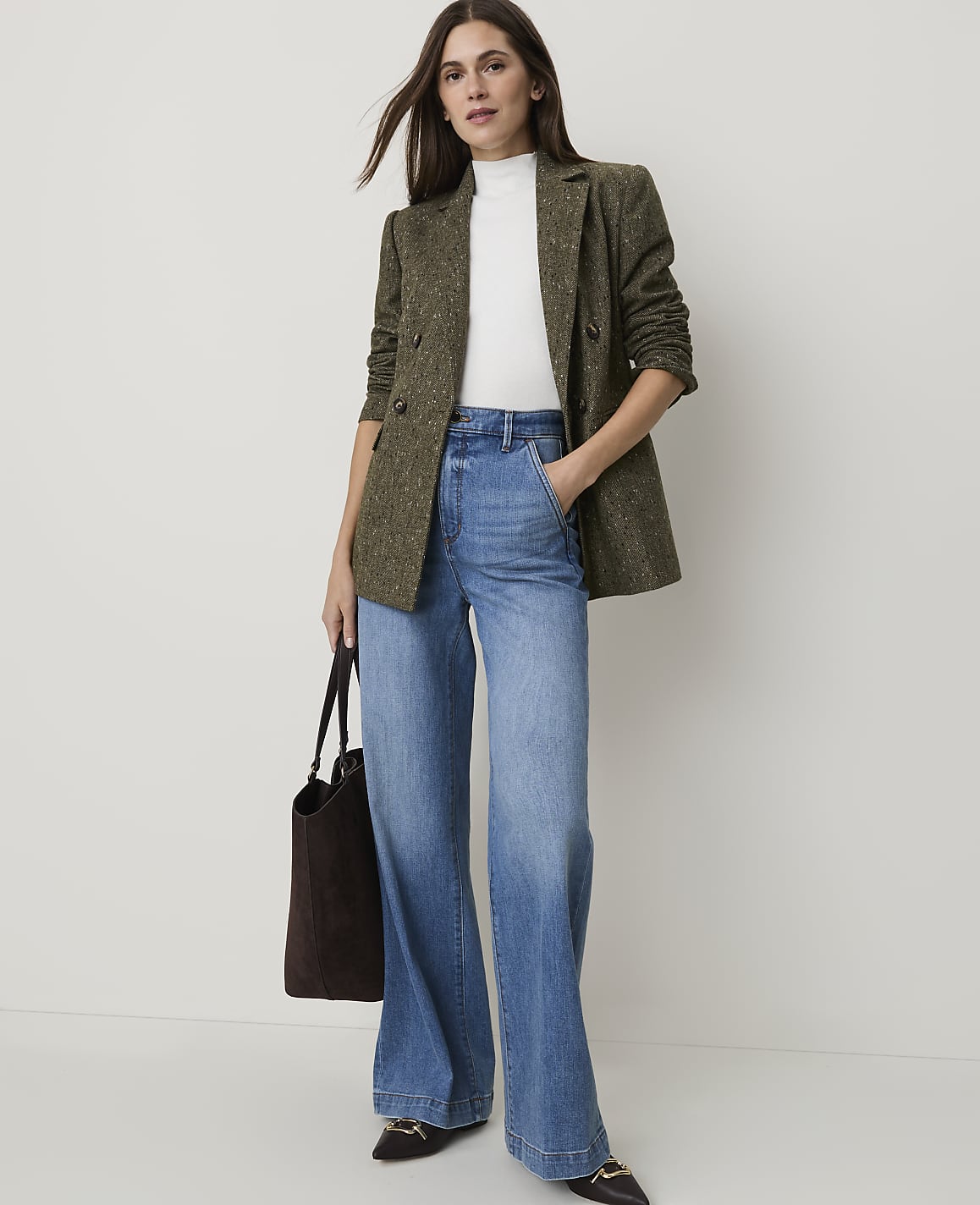 A woman in a tweed blazer and wide-leg jeans stands against a plain, light background, holding a dark tote bag.