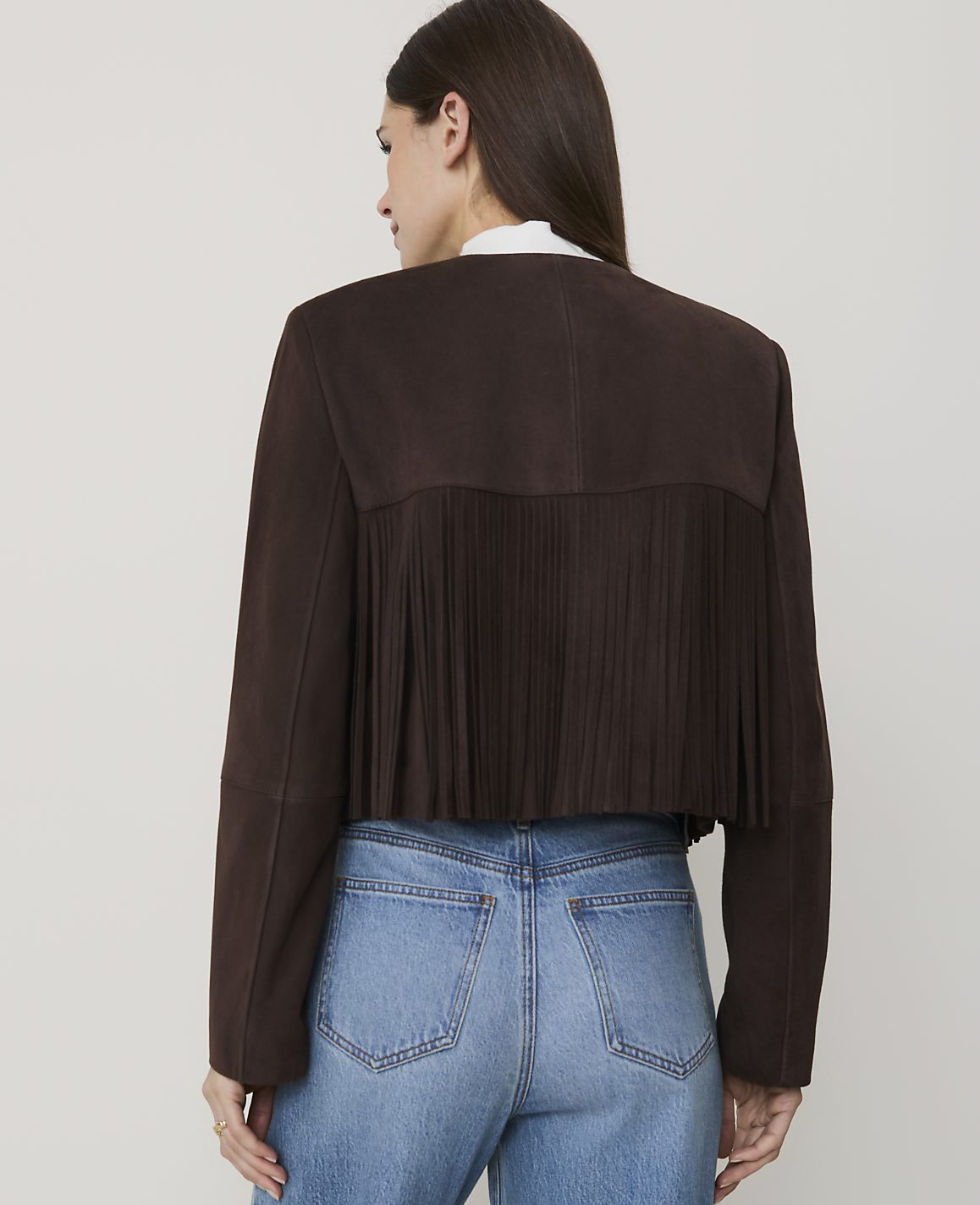 A woman in blue jeans and a brown suede jacket with fringe stands against a plain white background.