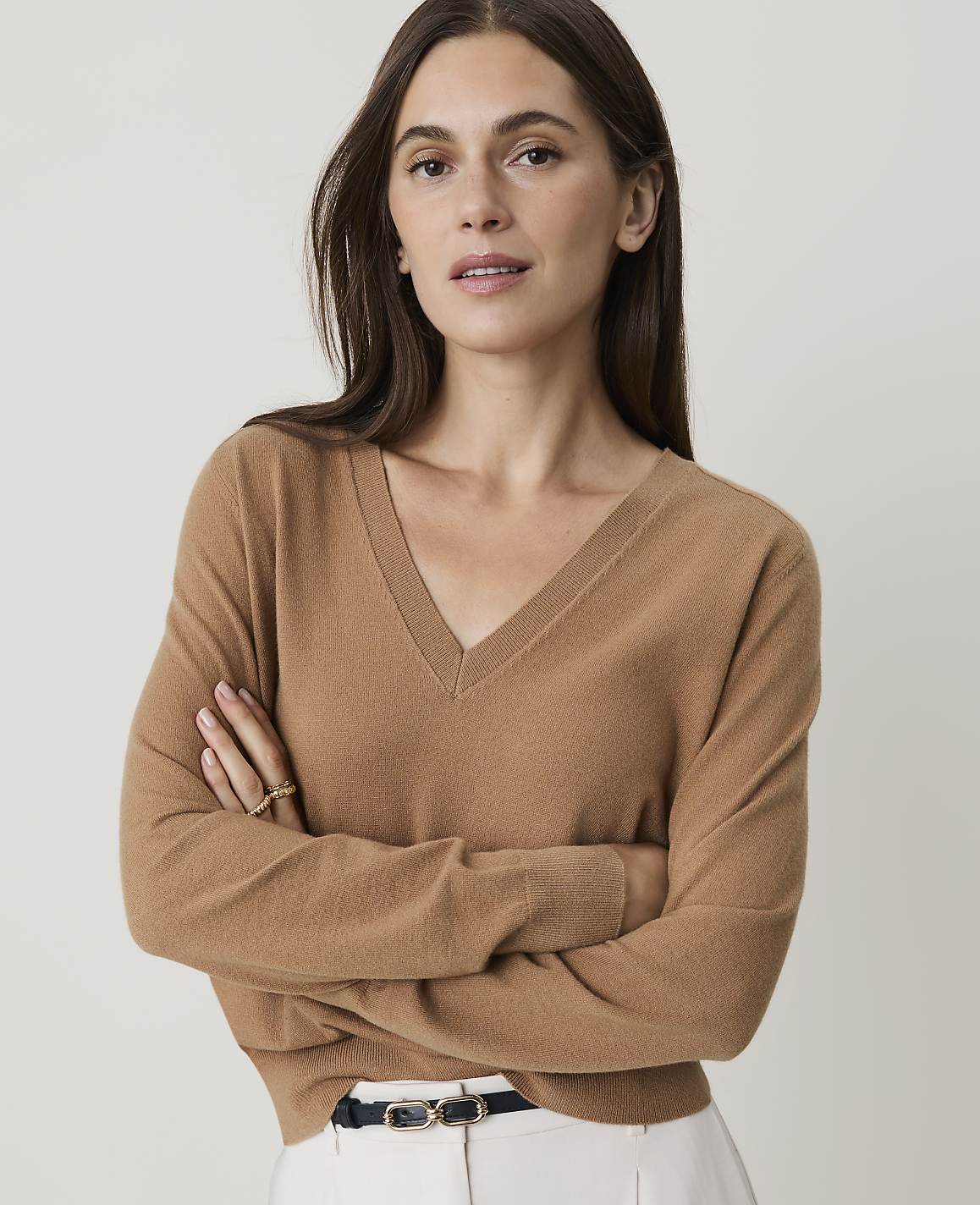 A woman in a tan v-neck sweater stands with her arms crossed against a plain white background.