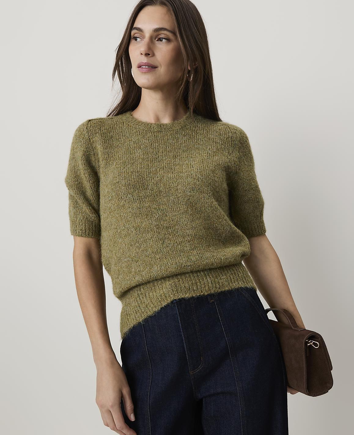 A woman in a green sweater and dark jeans holds a brown purse against a plain background.