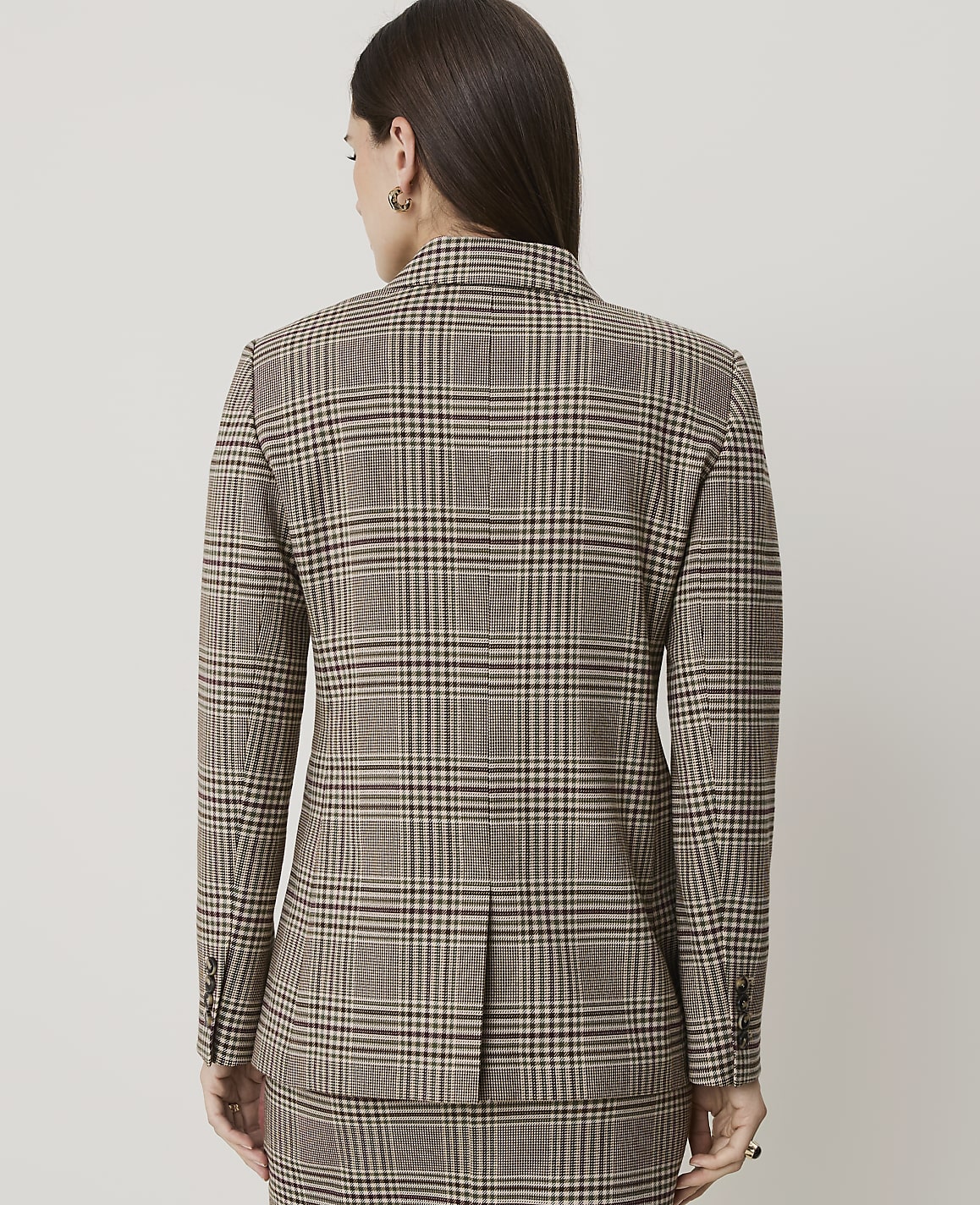 A woman in a brown plaid blazer and skirt stands against a plain, light gray background.