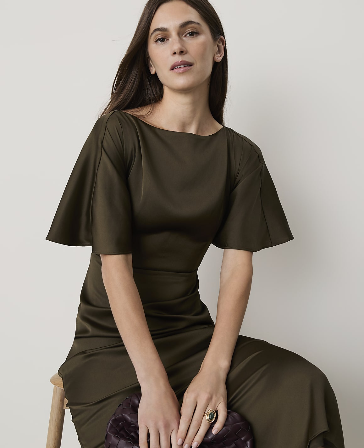 A woman in a flowing olive green dress sits on a stool in front of a plain white background.