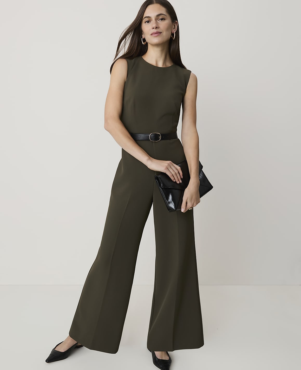 A woman in a green jumpsuit and black heels stands with a clutch in front of a plain white background.