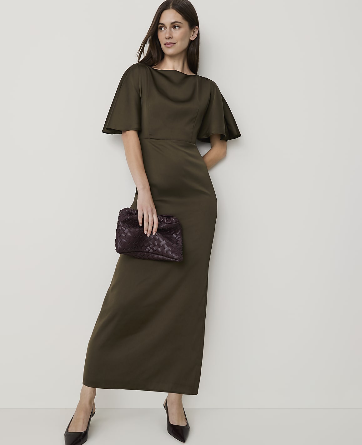A woman in a long, olive-green dress and dark heels holds a patterned clutch in front of a plain, off-white background.