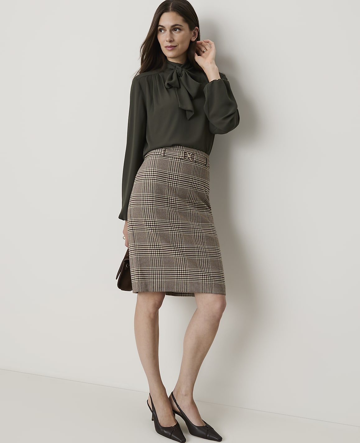 A woman in a dark green bow-neck blouse and a plaid pencil skirt stands against a plain, light-colored wall.