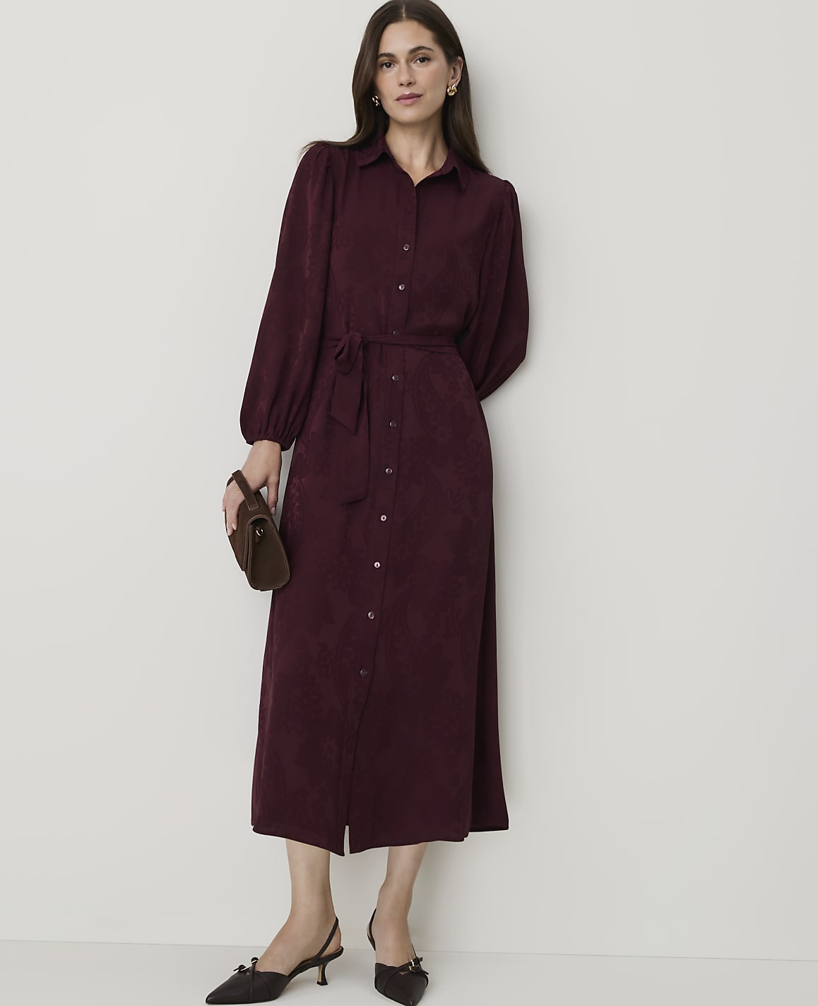 A woman in a burgundy shirtdress and pointed-toe heels stands against a plain, light-colored wall, holding a small clutch.