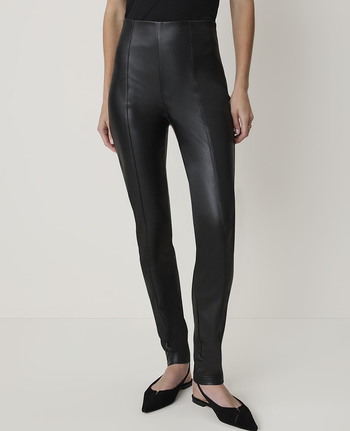 A person wearing black leather pants and black pointed-toe flats stands against a plain white background.