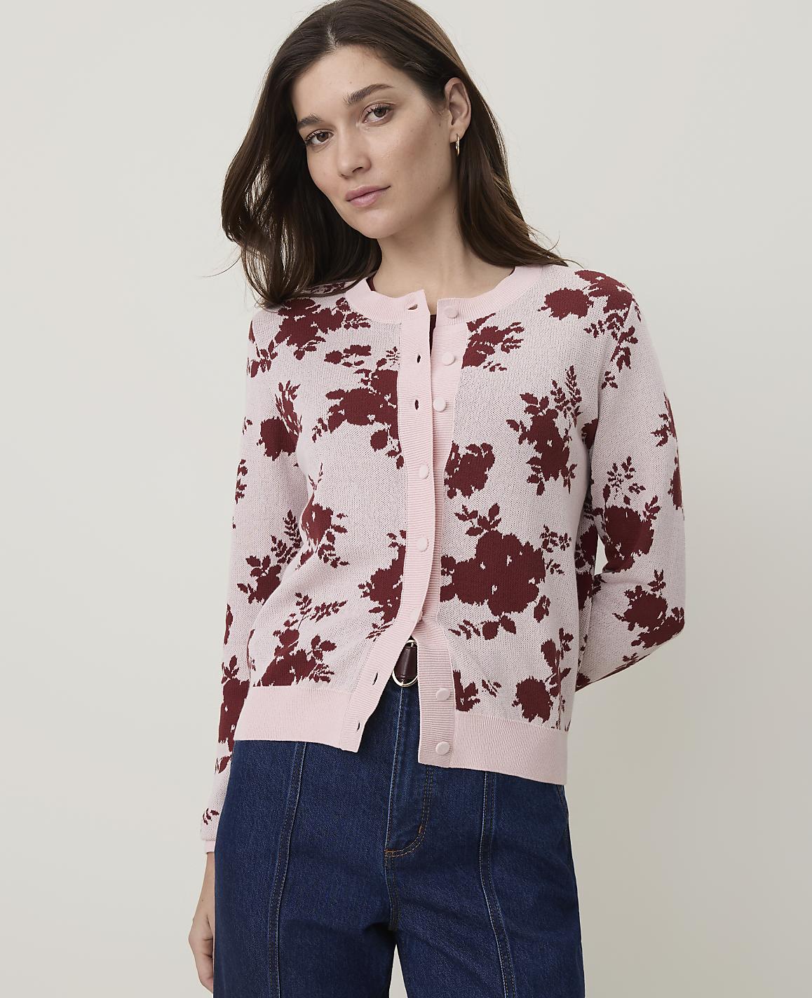 A woman in a pink floral cardigan and dark jeans stands against a plain background.