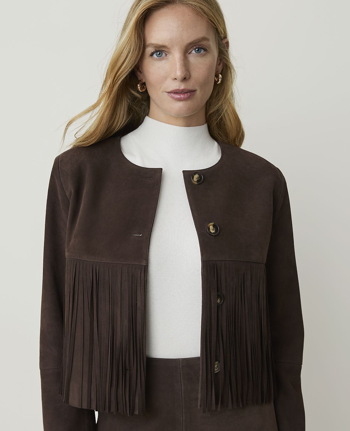 A woman with blonde hair and blue eyes, wearing a brown suede jacket with fringe, stands against a plain white background.