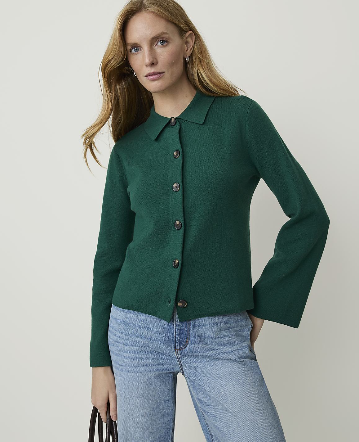 A woman in a green collared cardigan and blue jeans stands against a plain white background.