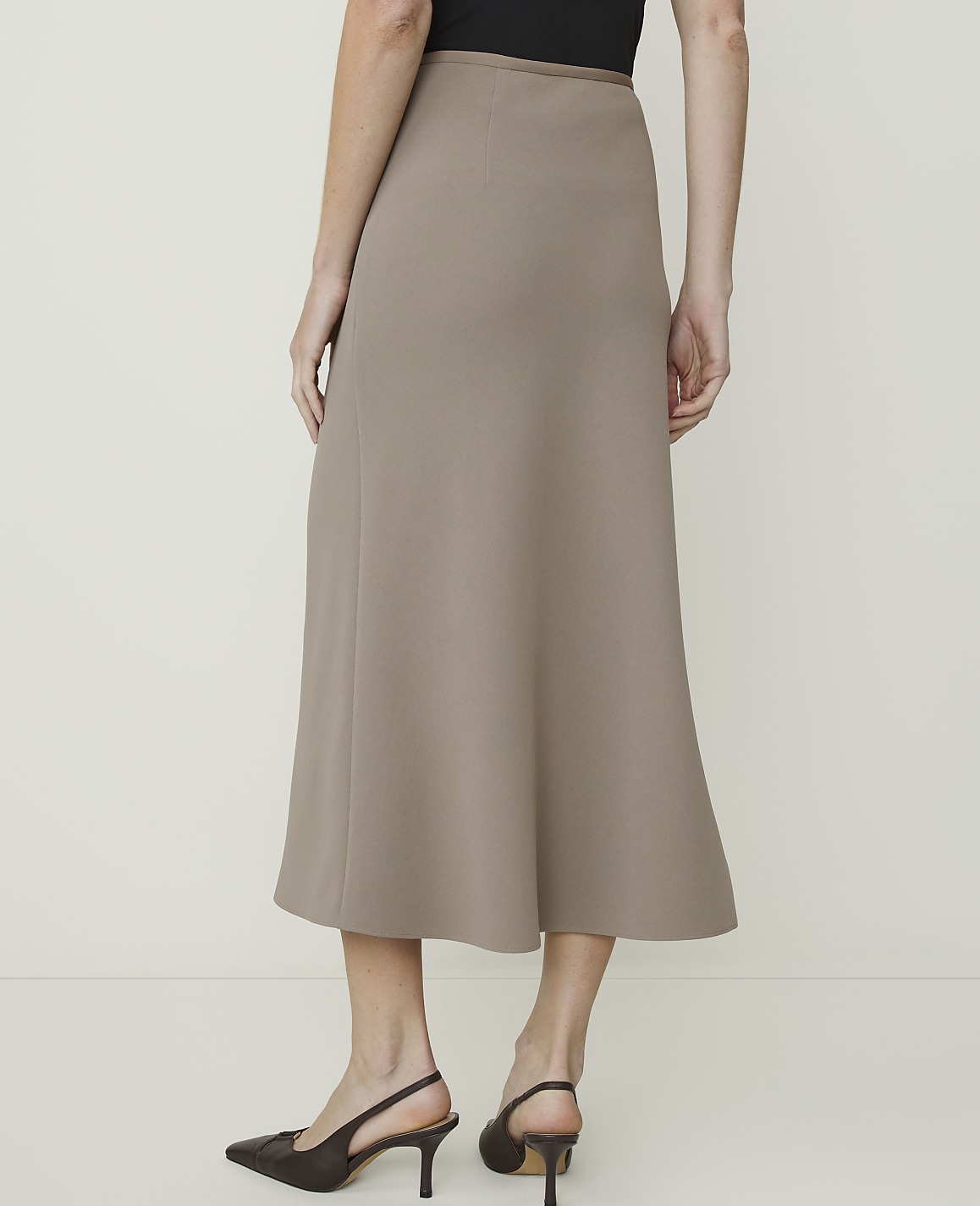 A woman in a taupe midi skirt and dark heels stands against a plain, light-colored background.