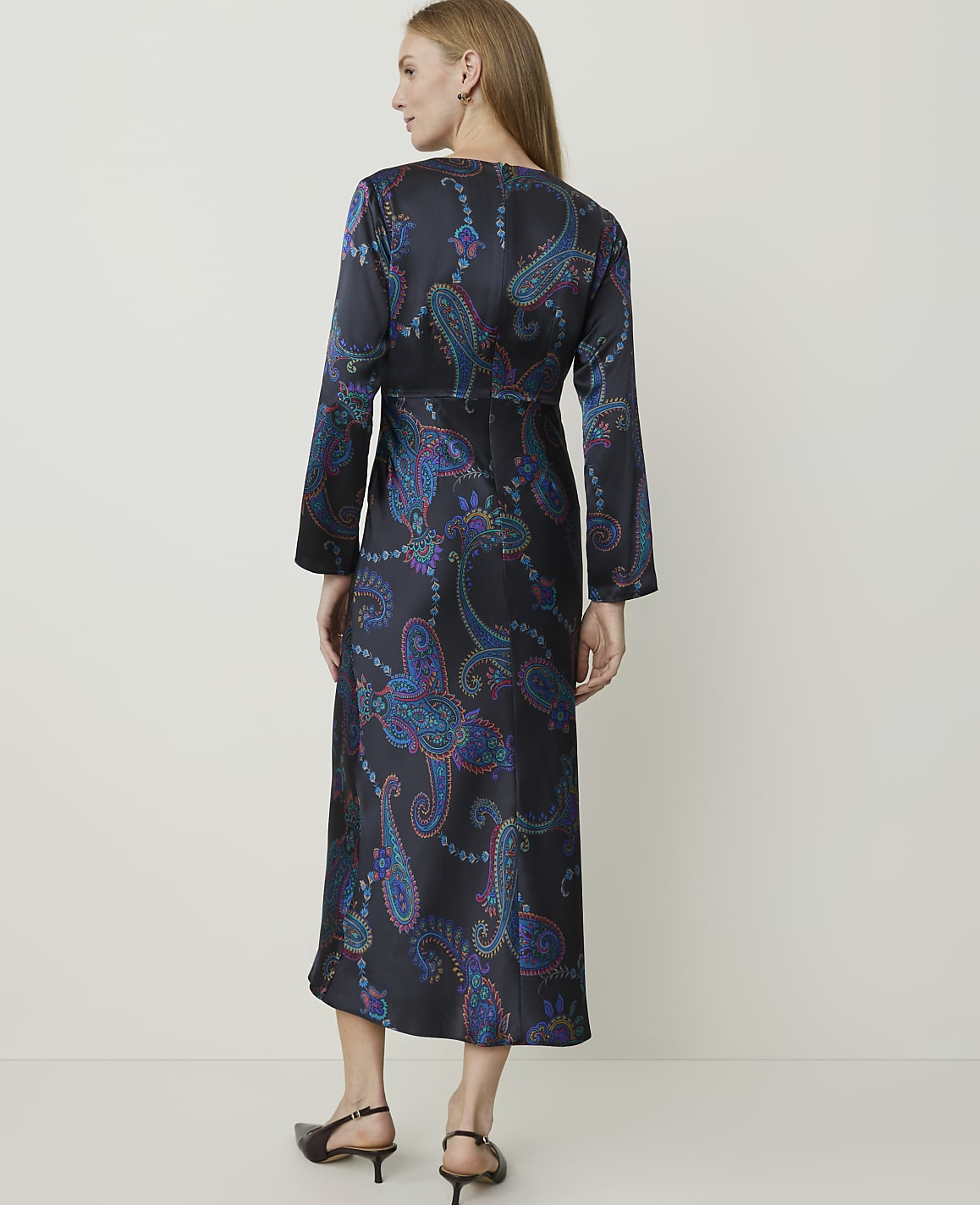 A woman in a long, dark, paisley-patterned dress stands against a plain, light-colored background.