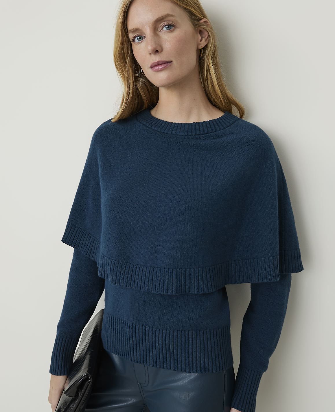 A woman in a teal sweater with a capelet and dark pants holds a black clutch against a plain background.