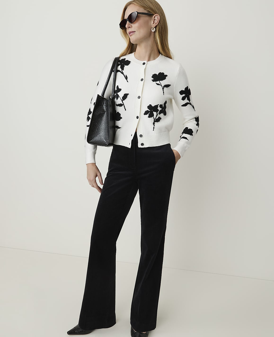 A woman in a floral cardigan and black flared pants stands against a plain white background.